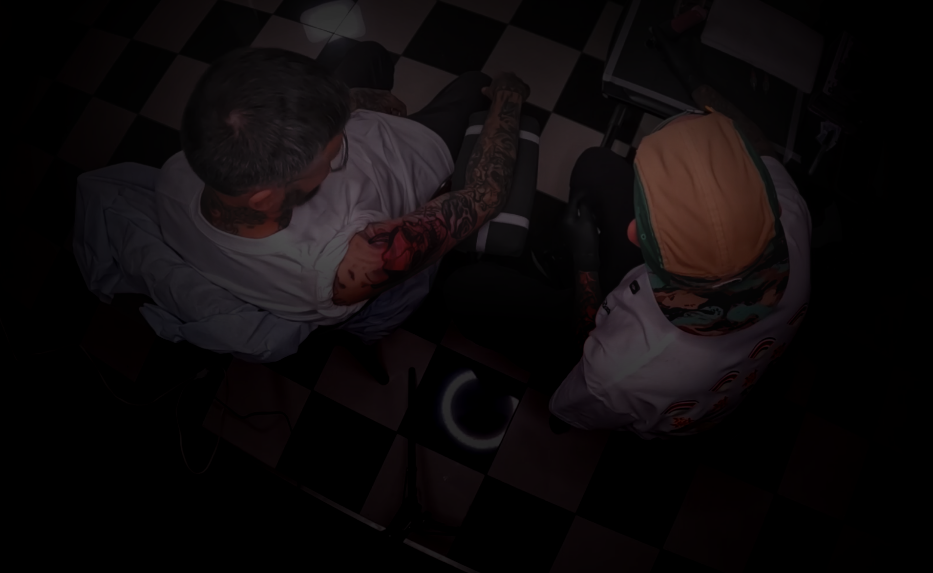 Two people sitting on a checkered floor, one with a tattoo on their arm.