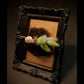 Decorative item with feathers and baby doll hand in a dark wooden frame on a brown background