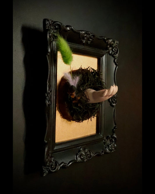  AN black frame featuring a nest and feathers, with a baby doll hand